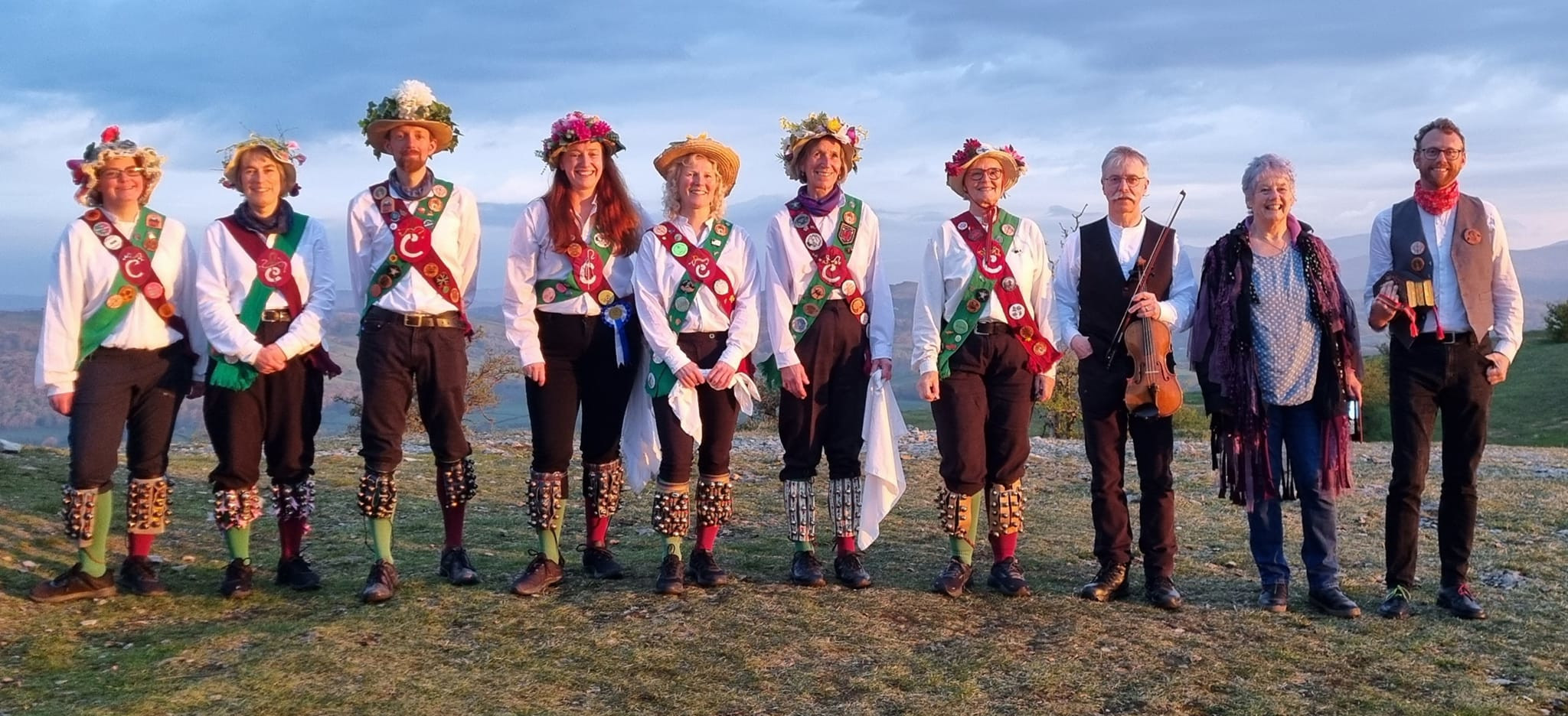 Eight jolly dancers in a line on a sunny day. They wear white shirts, black trousers, maroon and green baldricks and socks, and straw hats decorated with flowers.